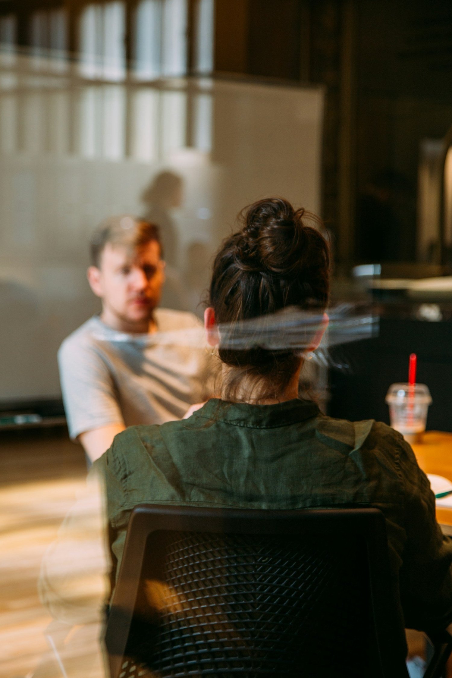 A woman with dark hair in a bun sitting at a table in front of a man with short hair, with a reflection of her in a glass window or partition. The woman's back is turned to the camera, and the man is looking towards her.
