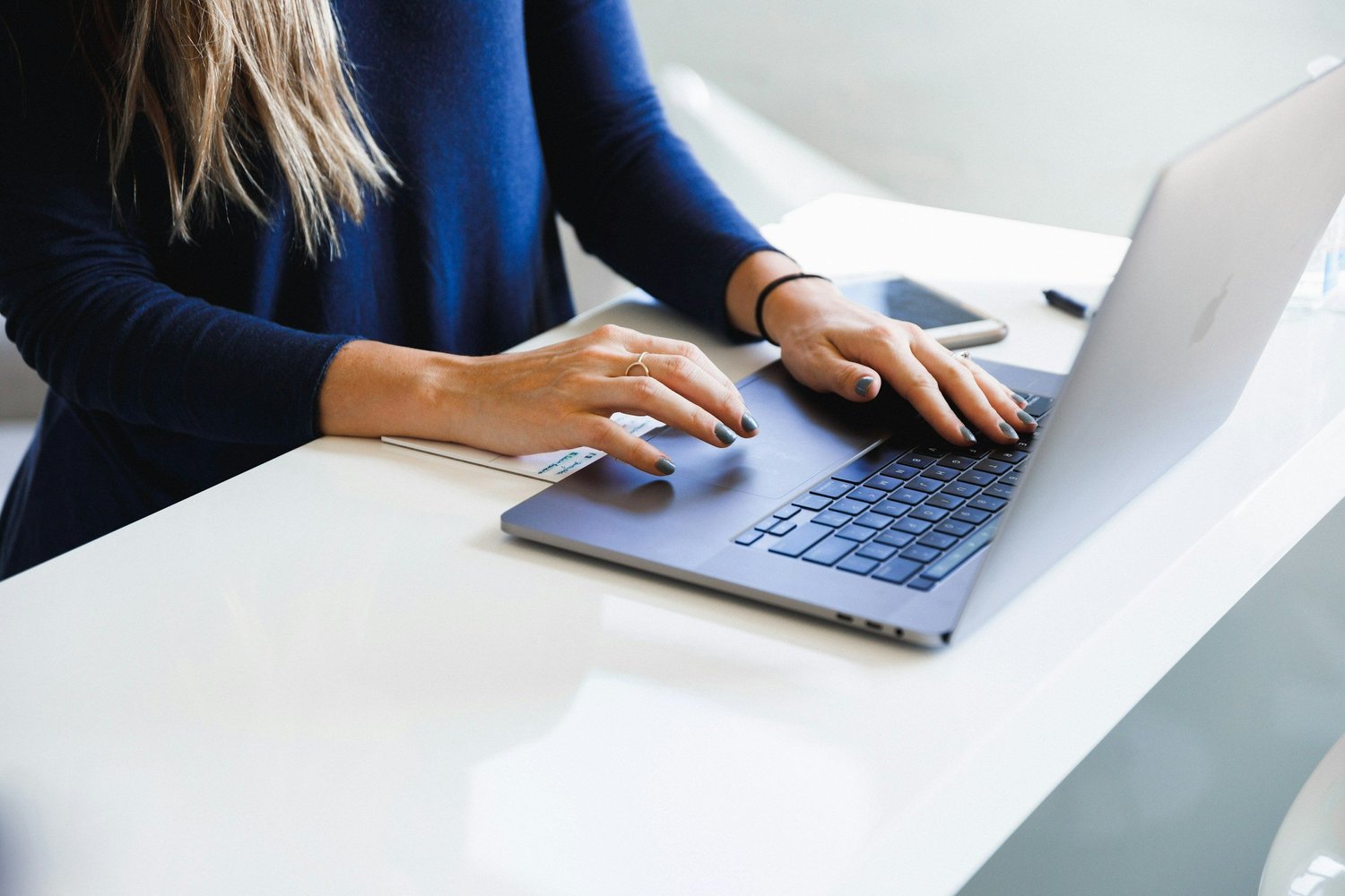 A woman working on a silver MacBook laptop at a white desk, with a smartphone and papers nearby.