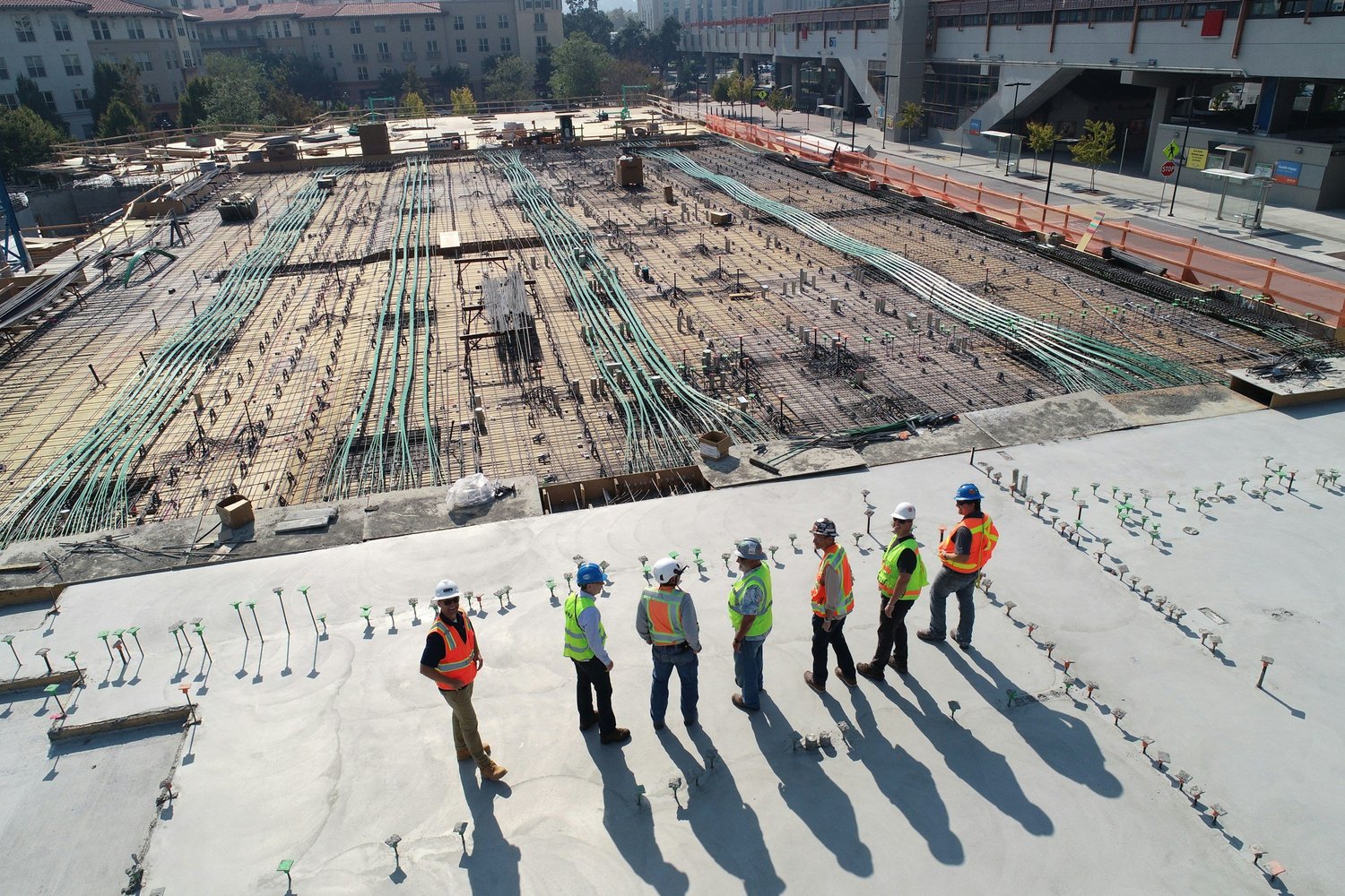 Construction workers in safety vests and helmets standing on a concrete floor at a construction site. In the background, the building's foundation and steel reinforcement bars are visible.