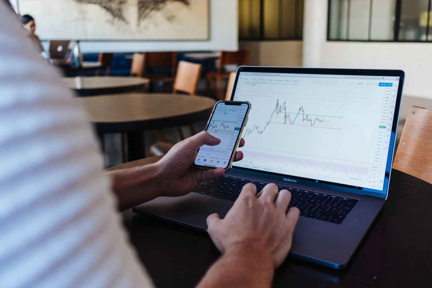 Person holding smartphone displaying financial charts next to a MacBook laptop showing similar financial charts on a table in a modern workspace.