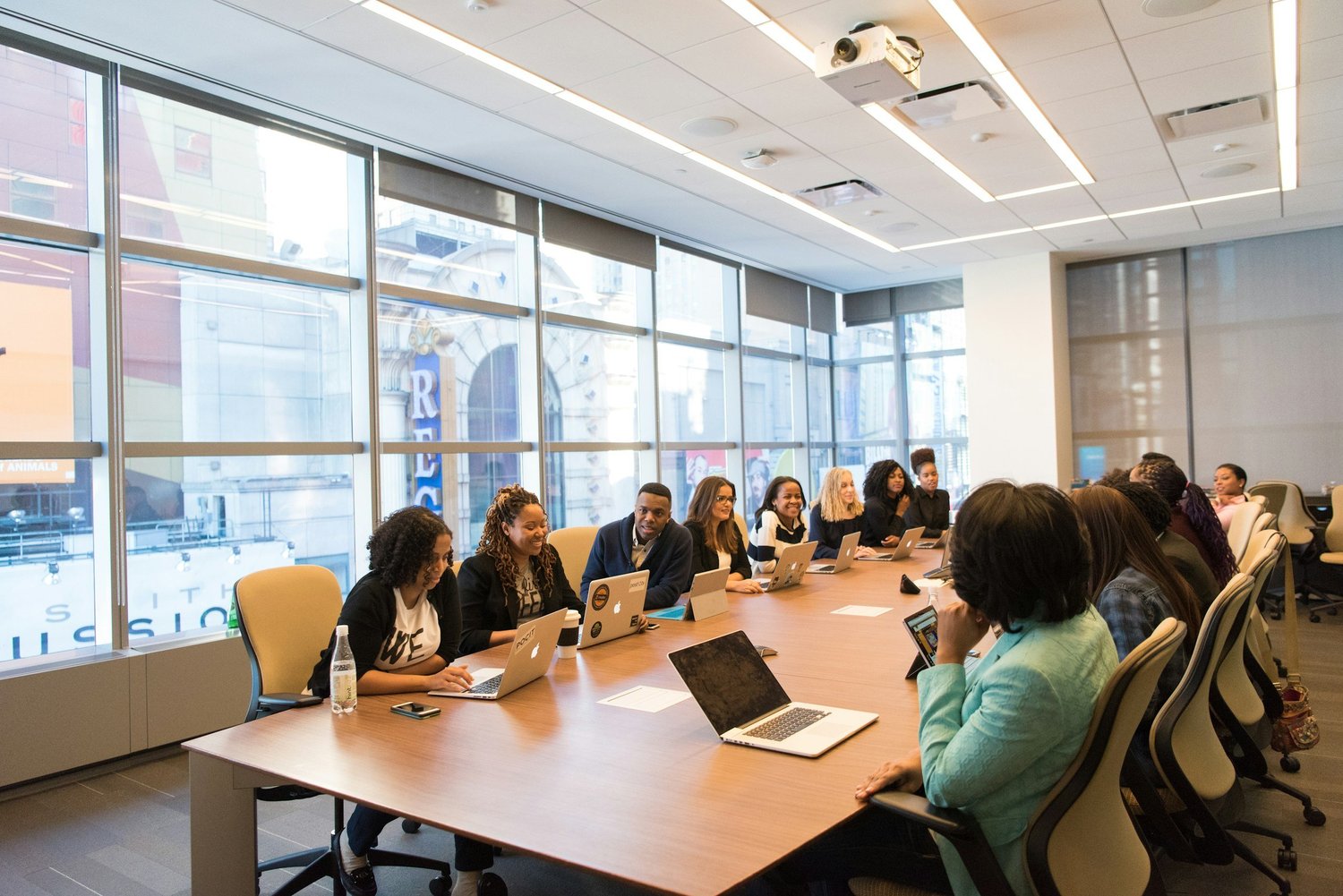 People seated around a large conference table in a modern office, many using laptops and tablets, with tall windows showing city buildings outside.