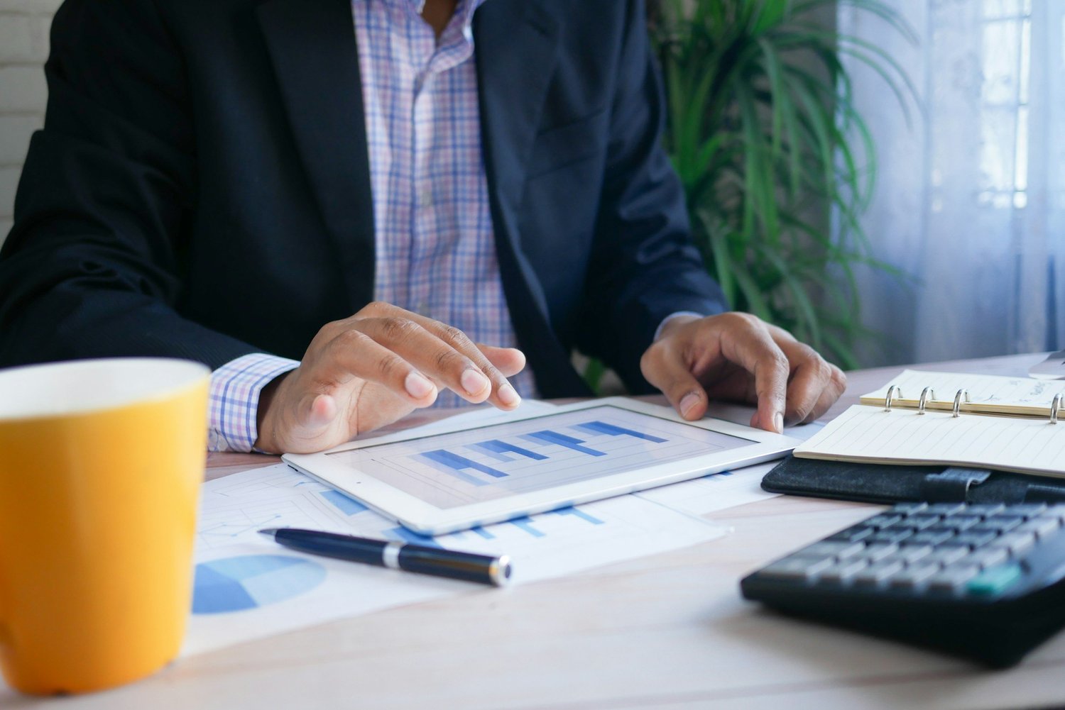 A person in a business suit works with documents, a tablet, and a calculator on a desk, with a coffee mug and notebook present. There are charts and graphs on the screen and papers.