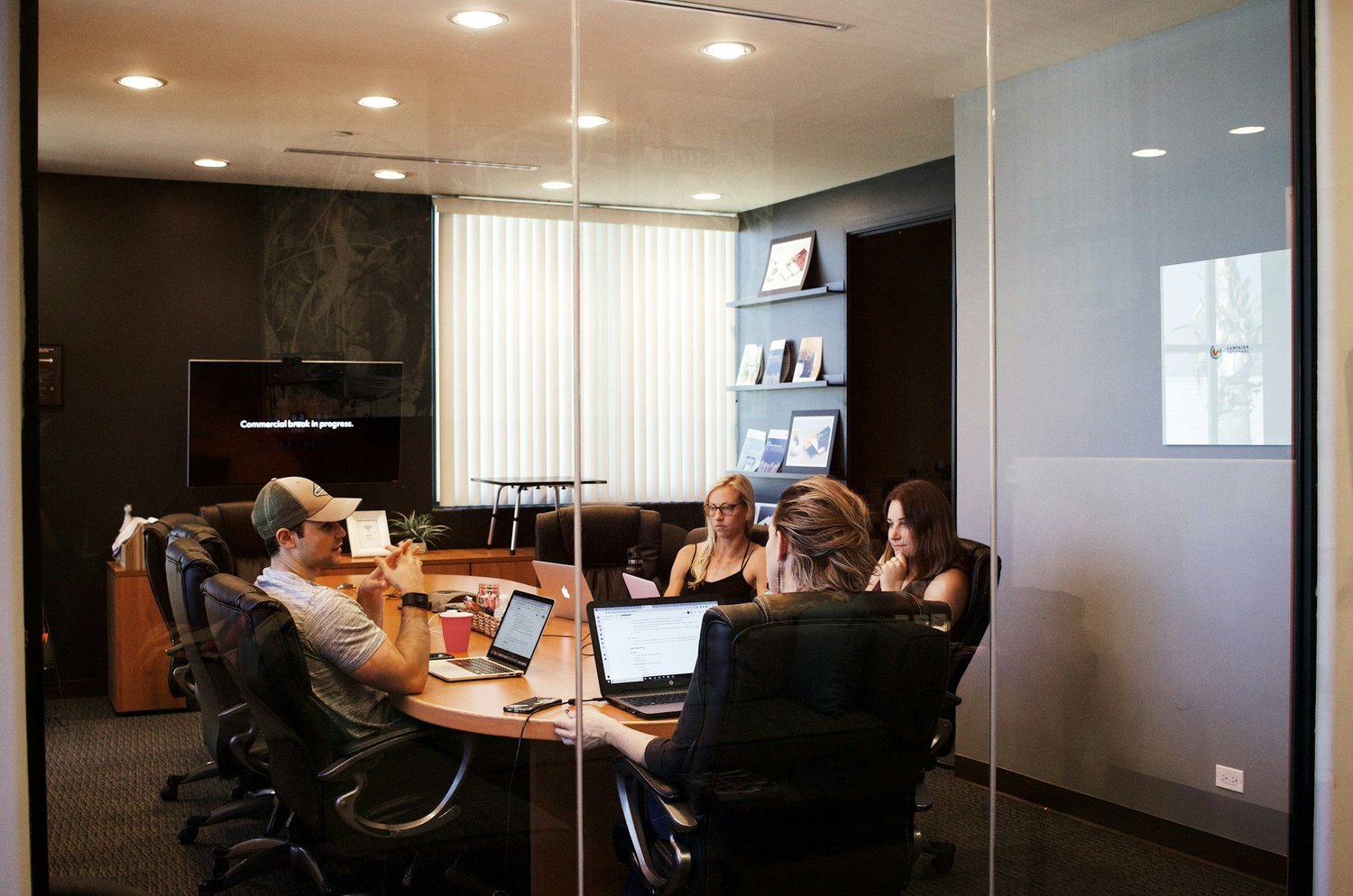 A group of five people having a meeting in a conference room seen through a glass wall. Four women and one man are sitting around a table with laptops and notebooks. The room has a television with a message, shelves with books or magazines, and a window with vertical blinds.