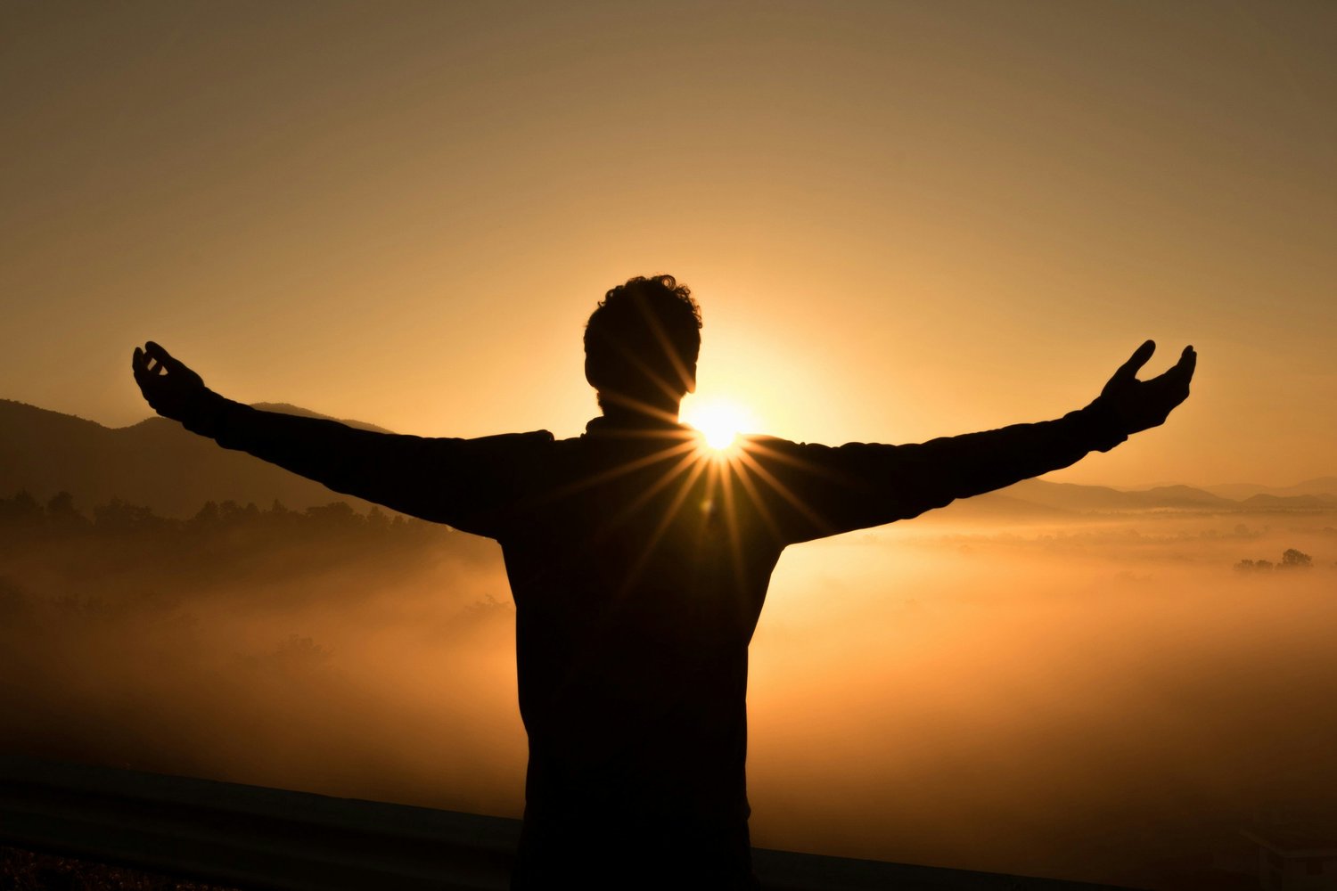 Silhouette of a person with arms outstretched at sunrise or sunset over a misty landscape.